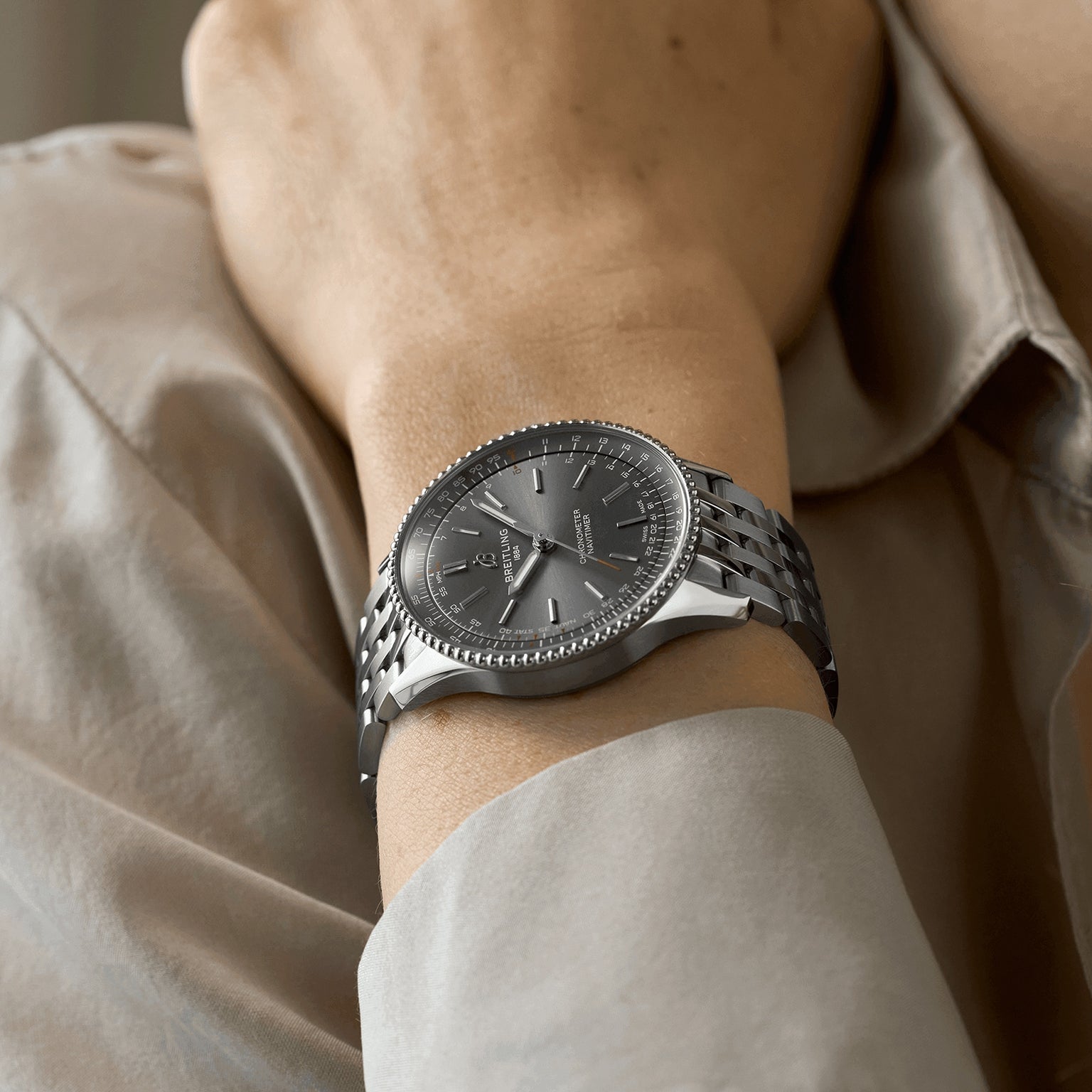 A person wearing a Breitling Navitimer Automatic 36 with a silver metal band and grey dial, paired with a beige long-sleeve shirt.