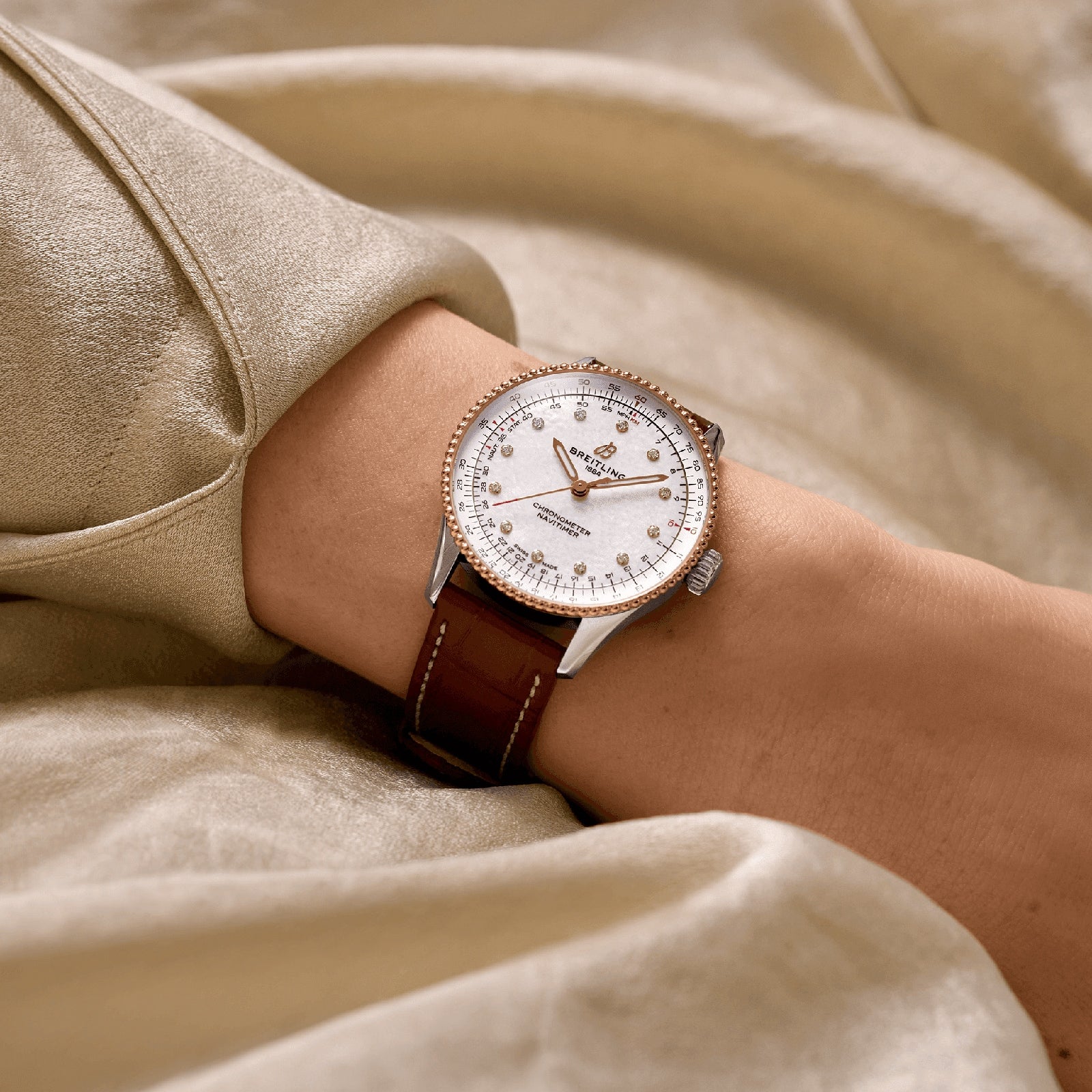 A person wears a Breitling Navitimer Automatic 36 with a mother-of-pearl dial, brown leather strap, and silver case, resting on champagne-colored satin fabric.