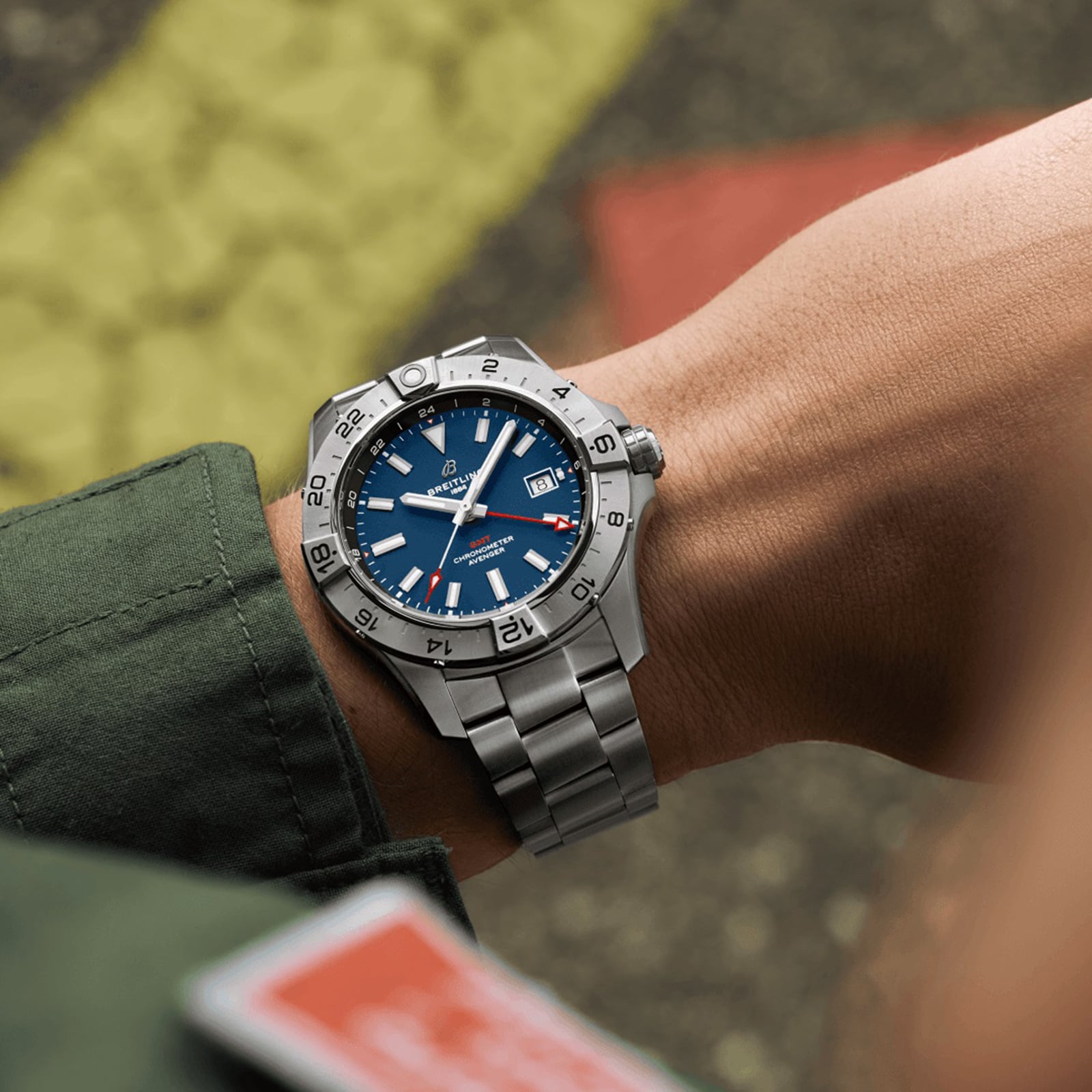 A person in a green jacket wears a Breitling Avenger Automatic GMT 44, featuring a stainless steel case, blue dial, date display, 24-hour bezel, and water resistance.
