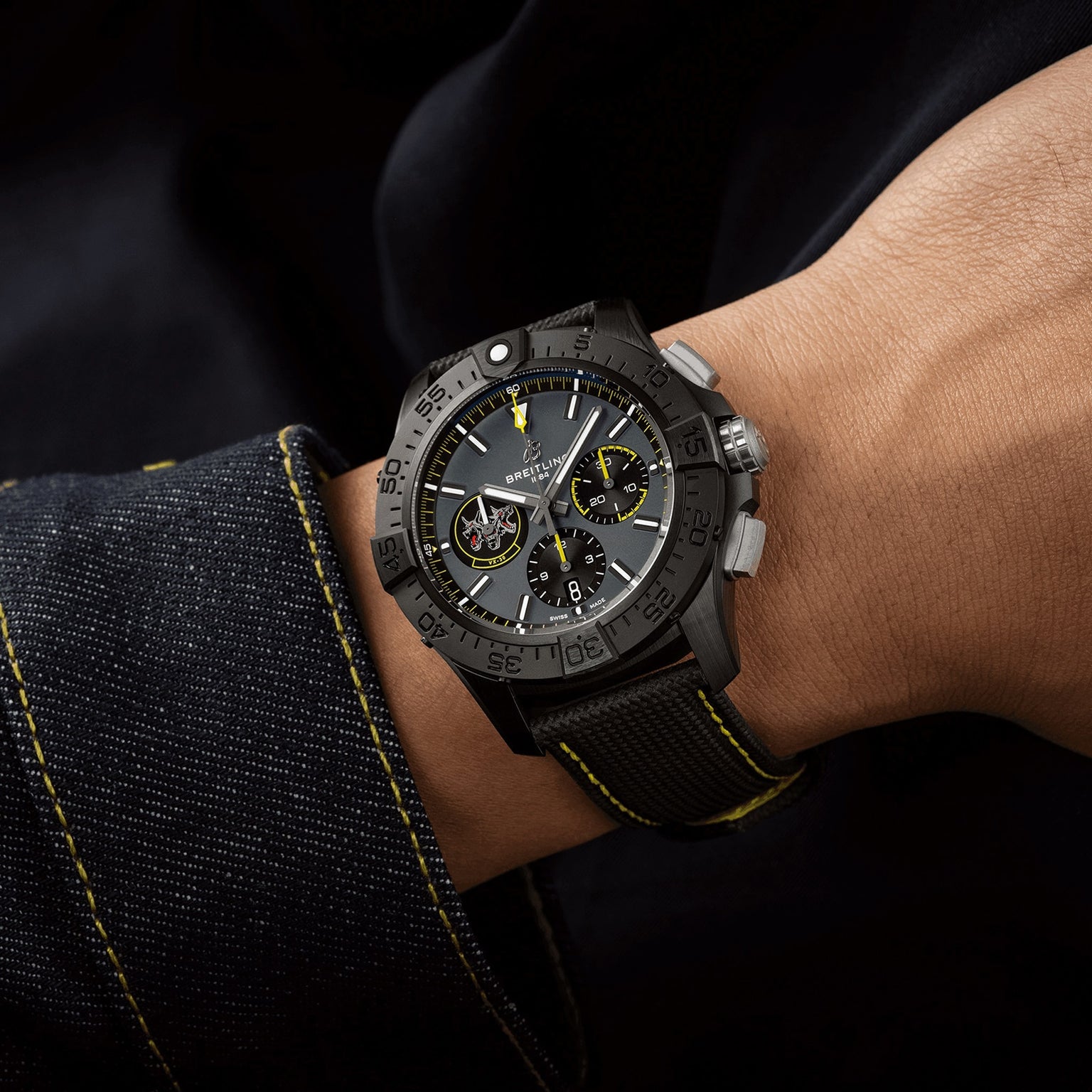 A person sports the Breitling Avenger B01 Chronograph 44 Night Mission Bloodhounds, a black watch with yellow stitching, paired with a dark outfit featuring matching cuffs—evoking jet pilot precision.