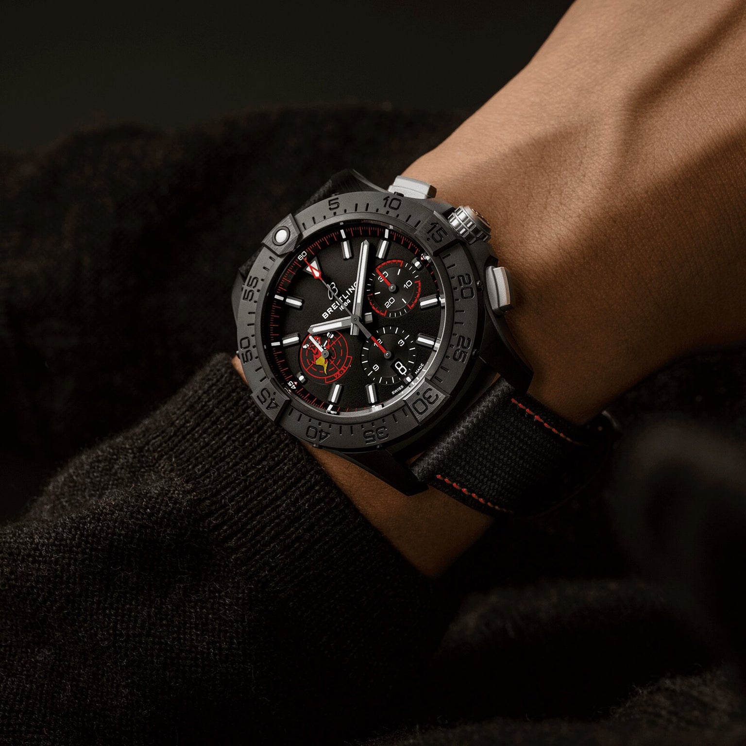 A Breitling Avenger B01 Chronograph 44 Night Mission Dust Devils wristwatch with a black fabric strap and red stitching is worn, showing the time and two subdials on a dark background, powered by the Breitling Manufacture Caliber 01.