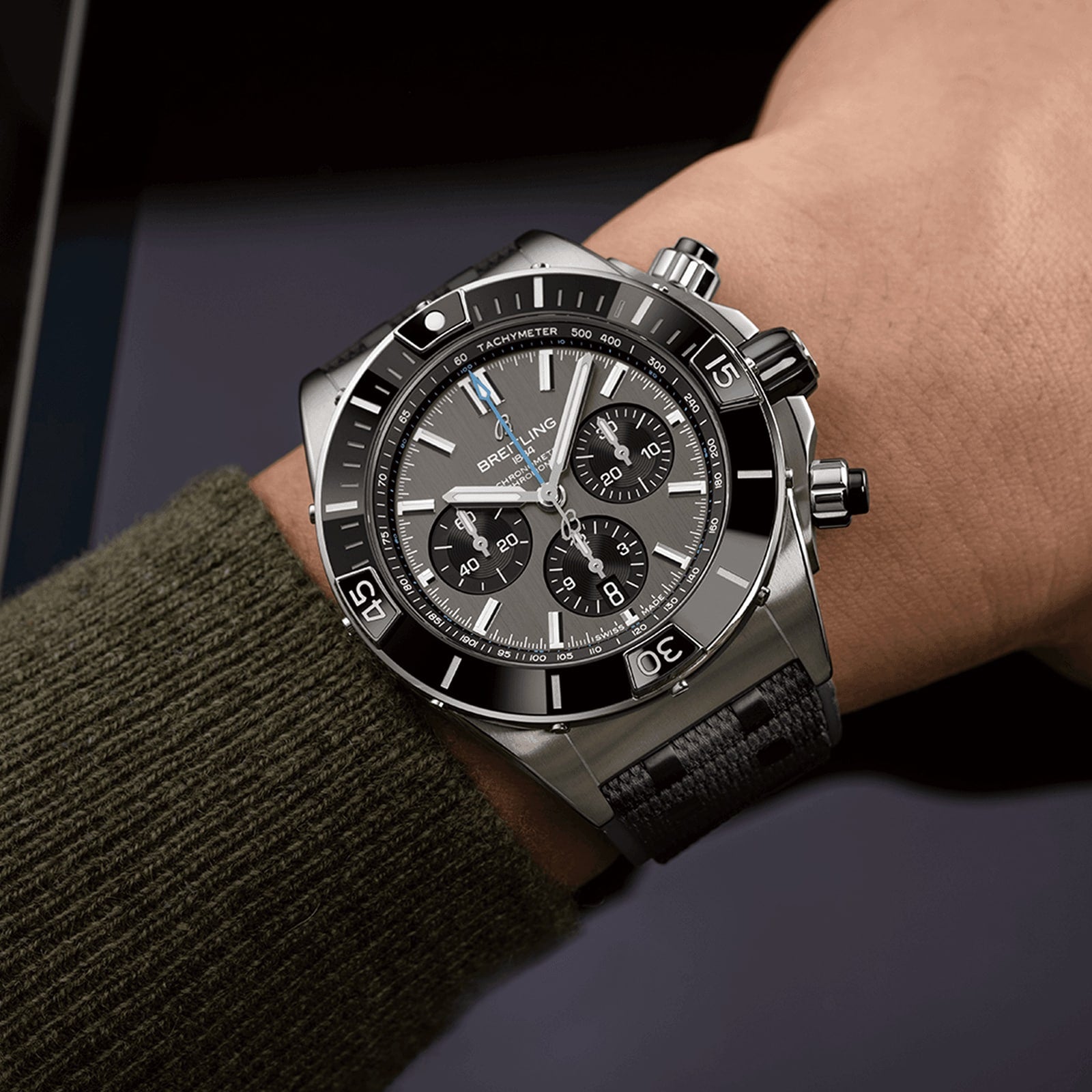 A person wearing a Breitling Super Chronomat B01 44, a black and silver chronograph wristwatch with a tachymeter, multiple dials, and a black strap, shown close-up on the wrist.