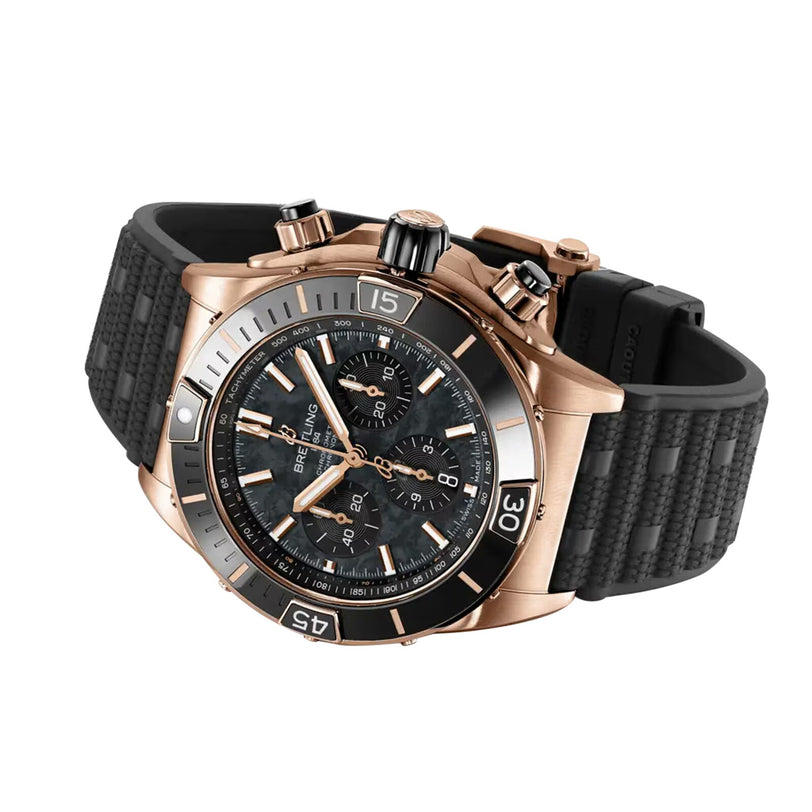 A Breitling Super Chronomat B01 44 wristwatch in gold tone with a black textured rubber strap, black bezel, and three subdials is displayed at an angle on a white background. This COSC-certified chronometer blends precision and bold style.