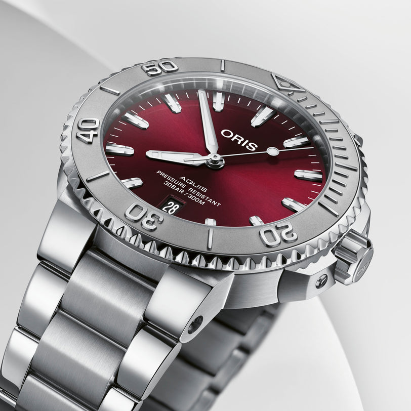 A close-up of the Oris Aquis Date Relief, a stainless steel wristwatch with a red dial, silver bezel, time set to 10:10, and a date display at the 6 o’clock position.