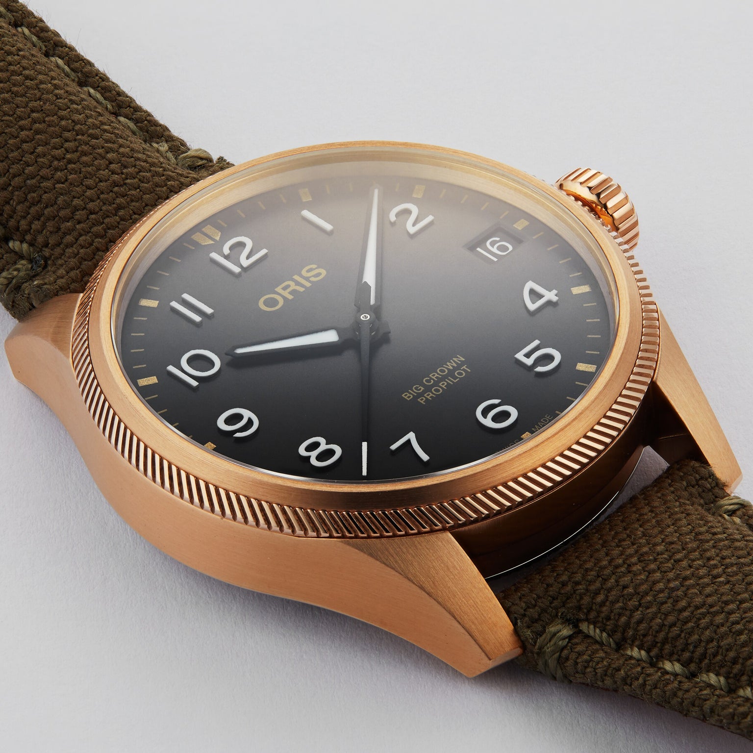 The Oris Big Crown ProPilot Big Date wristwatch features a bronze case, black dial with white numerals, Swiss mechanical movement, date display at 3 o’clock, and an olive green fabric strap.