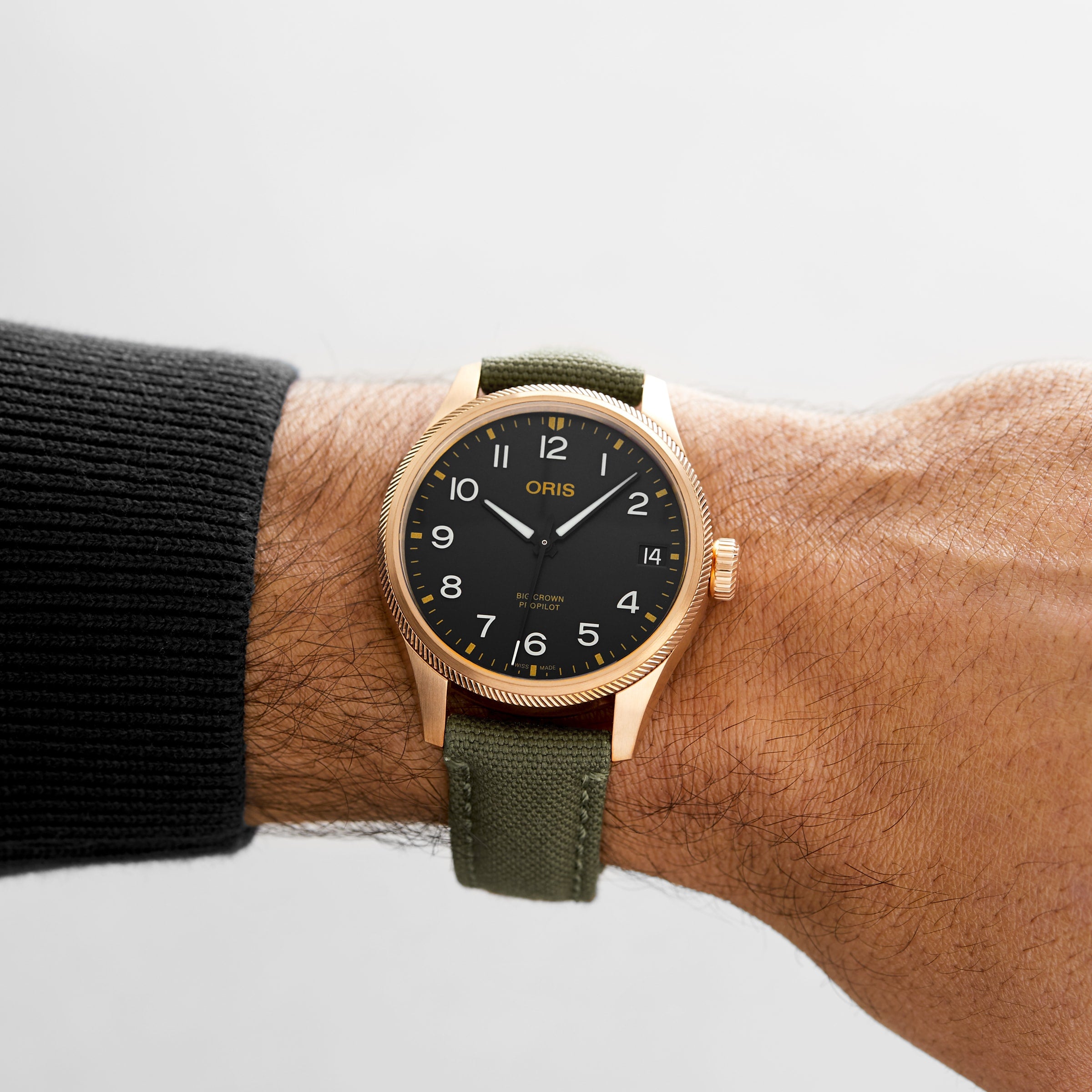 A person wearing an Oris Big Crown ProPilot Big Date wristwatch with a black dial, gold bezel, green fabric strap, and Swiss Made mechanical movement on their left wrist.