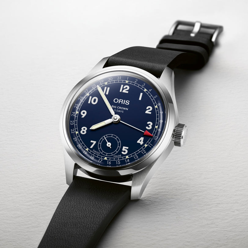 The Oris Big Crown Pointer Date Calibre 403 features a stainless steel case, blue dial, black leather strap, white numerals, and a red-tipped date hand. Shown photographed on a light-colored surface.