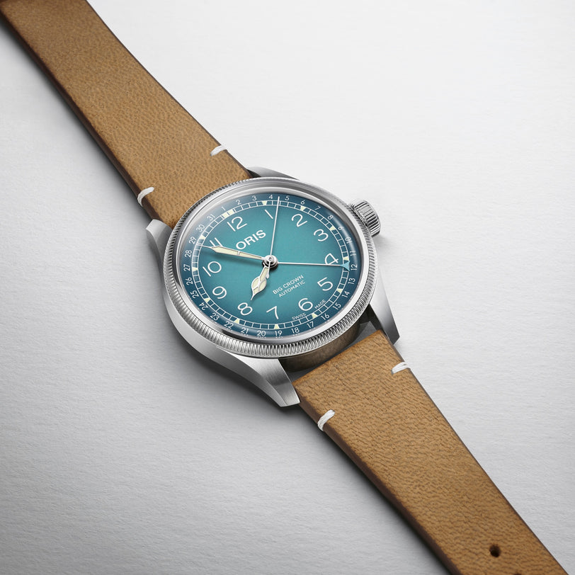 A silver Oris Big Crown Oris X Cervo Volante watch with a blue dial, white numerals, and a tan leather strap is displayed on a light gray surface.