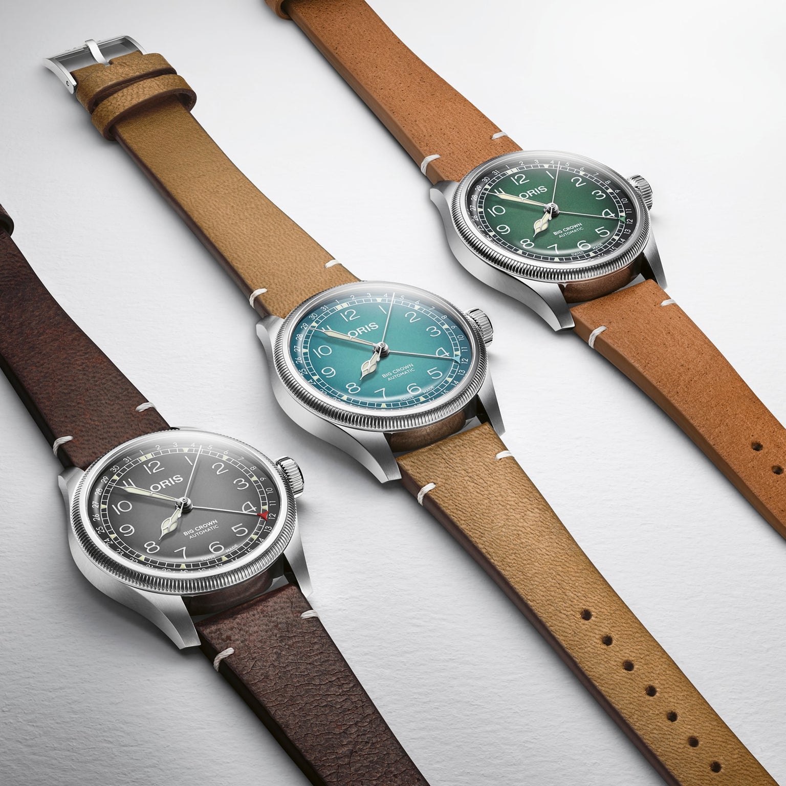 Three Oris Big Crown Oris X Cervo Volante watches with leather straps are shown side by side, each with a stainless steel case, large numerals, and gray, teal, or green dials that showcase the classic style of the series.