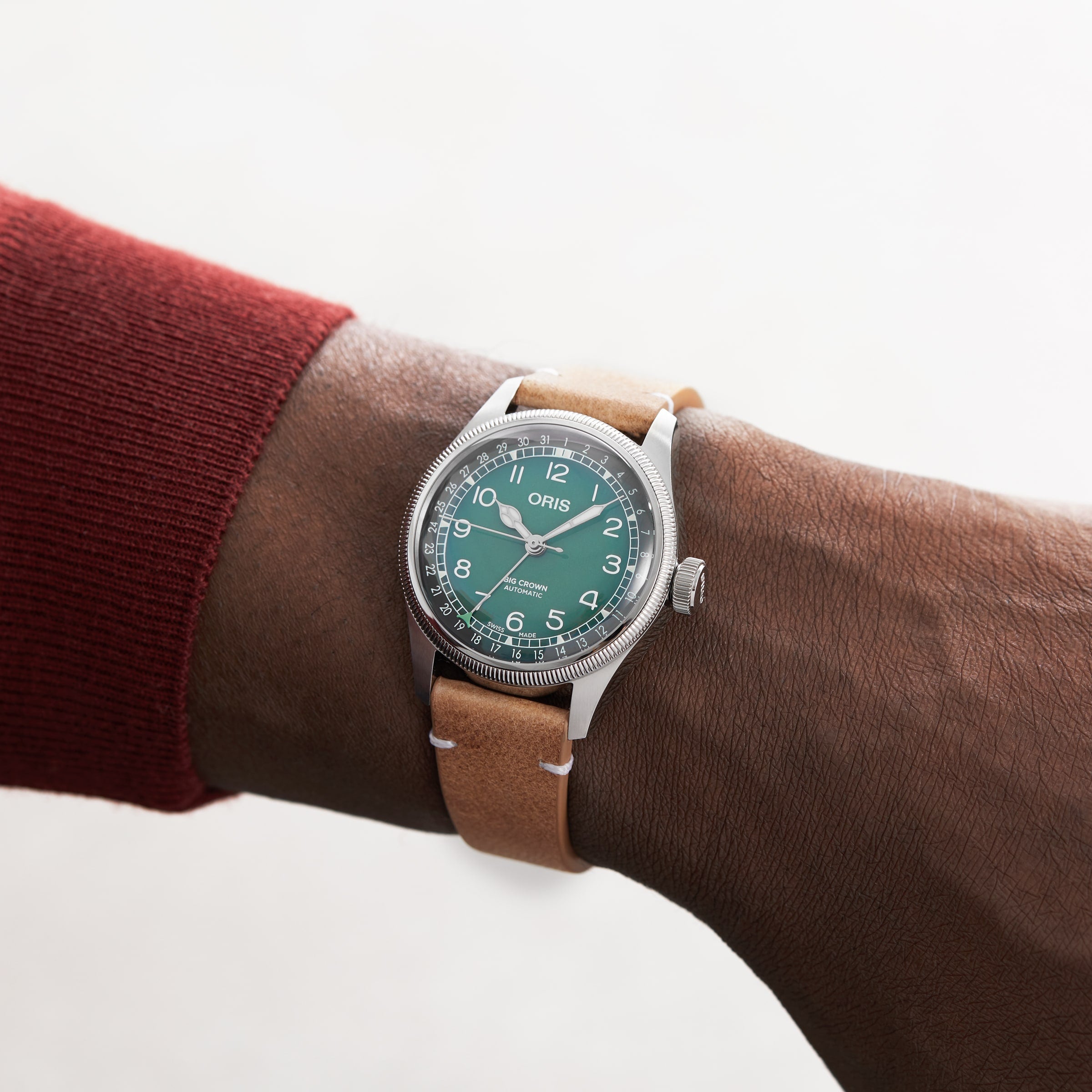 A person wearing the Oris Big Crown X Cervo Volante watch, featuring a green dial with white numerals and a tan leather strap. The Oris brand and classic ProPilot-inspired design are visible on the face.