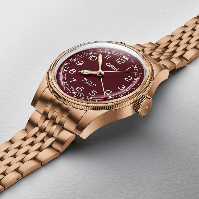 The Oris Big Crown Bronze Pointer Date wristwatch features a red dial and gold bracelet, shown on a light gray textured surface.