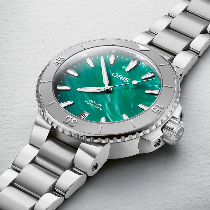 Close-up of the Oris Aquis Oris X Bracenet watch with a green mother-of-pearl dial, Super-LumiNova silver hour markers, and a stainless steel bracelet on a light textured surface.