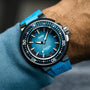 A close-up of a wrist wearing the Oris Aquis Pro 4000M watch, featuring a blue wave-patterned dial, bold white markers, blue rubber strap, and Oris’s trademark Rotation Safety System.