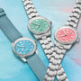 Three Oris Divers Sixty‑Five Cotton Candy watches with blue, green, and pink dials are displayed on a pastel background; one features a fabric strap.