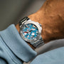 An Oris Aquis Date Upcycle Calibre 400 with a blue and white camo-patterned dial is worn with a light blue long-sleeve shirt, reflecting Oriss dedication to mechanical watchmaking and sustainability.