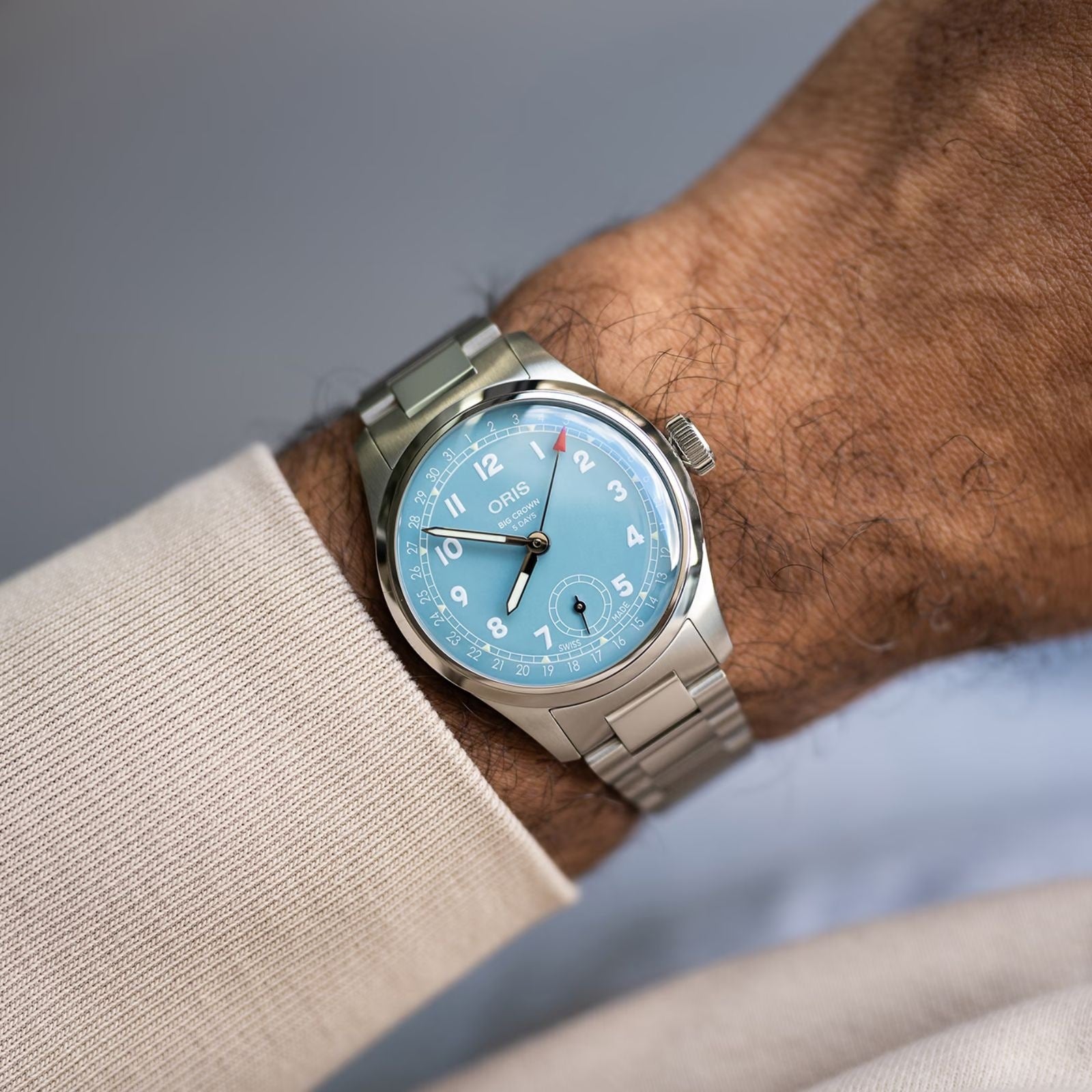 A wrist wears the Oris Big Crown Pointer Date Calibre 473, featuring a silver case, light teal blue dial, silver hour markers, and a metal band, set against a neutral background.