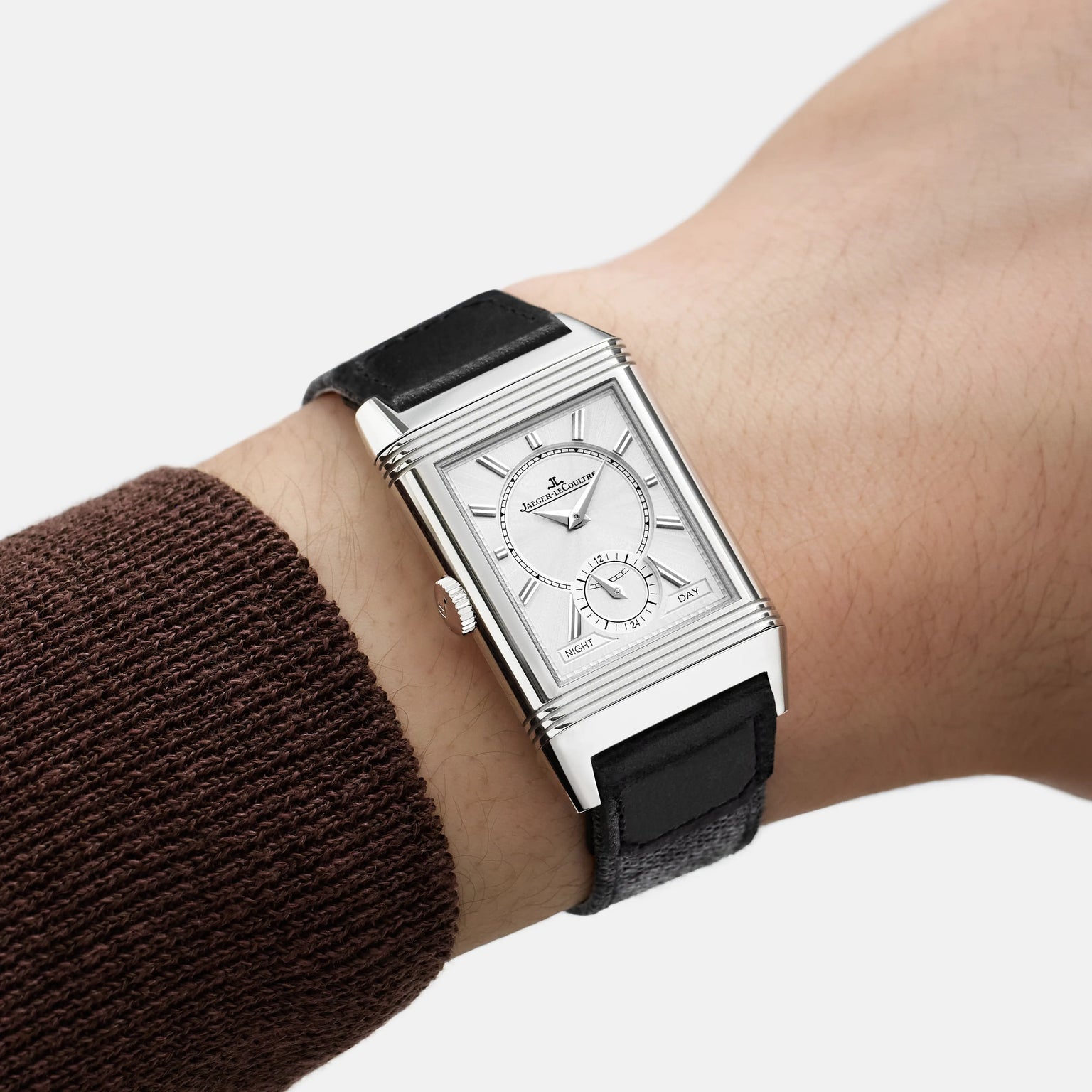 A Jaeger-LeCoultre Reverso Tribute Duoface Small Seconds, a rectangular steel wristwatch with a white dial, small seconds feature, and black leather strap, is worn on the wrist of someone in a brown sweater.