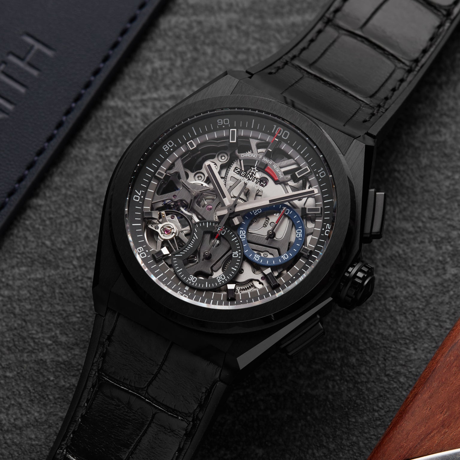 The Zenith Defy El Primero 21, a black luxury skeleton watch with visible mechanical movement, high-frequency chronograph, black leather strap, and blue accents, rests on a dark textured surface near leather, exuding urban sophistication.