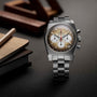 The Zenith Chronomaster Revival El Primero A384, a stainless steel chronograph with a brown gradient dial, three white subdials, and a tonneau case, is shown on a dark surface beside notebooks and wooden objects.