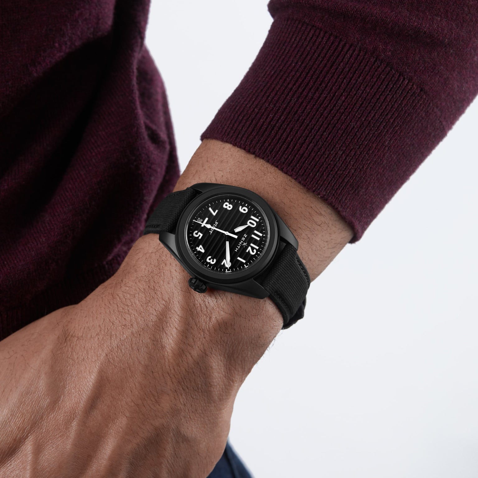 A close-up of a wrist wearing the Zenith Pilot Automatic, featuring a black strap and white numerals, set against a maroon long-sleeve shirt.