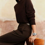 A person in a dark brown sweater and pants sits on a wooden chair, wearing the Zenith Chronomaster Original watch with a chronograph movement on their left wrist. Their head is not visible.