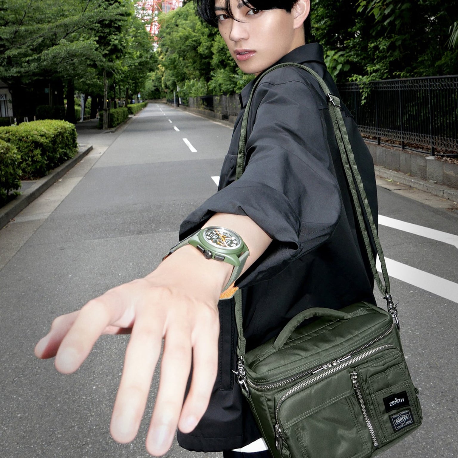 A person in a black jacket and green bag extends their arm, displaying the Zenith Limited Edition Pilot Big Date Flyback Porter with a khaki-green ceramic case, on an empty street lined with greenery.
