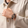 A person in a beige jacket is wearing a Zenith Chronomaster Original, a black and silver watch with a black strap, on their left wrist.