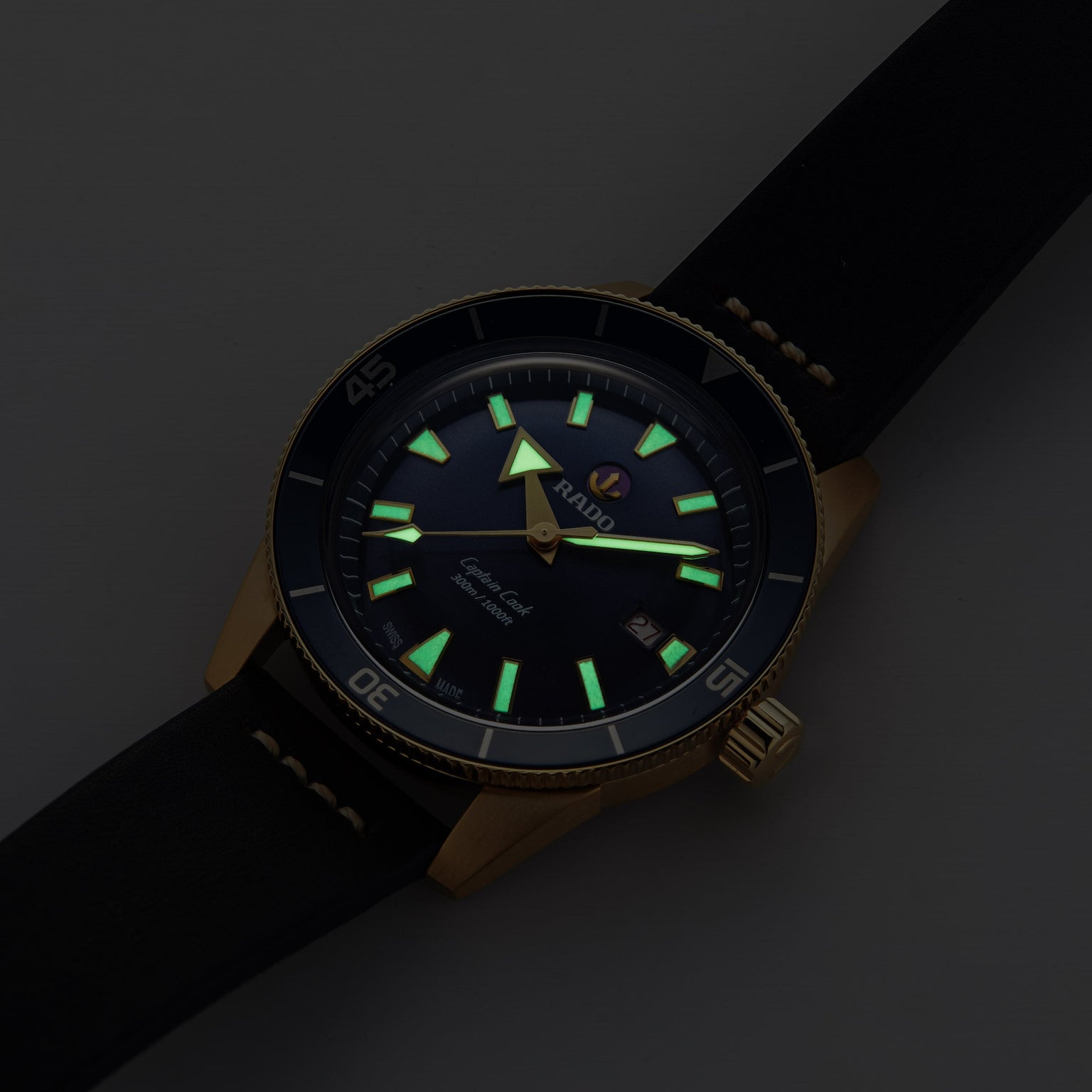 The Rado Captain Cook Automatic Bronze wristwatch features a black dial, bronze case, and black strap with luminous hour markers and hands visible in low light.