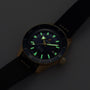 The Rado Captain Cook Automatic Bronze wristwatch features a black dial, bronze case, and black strap with luminous hour markers and hands visible in low light.