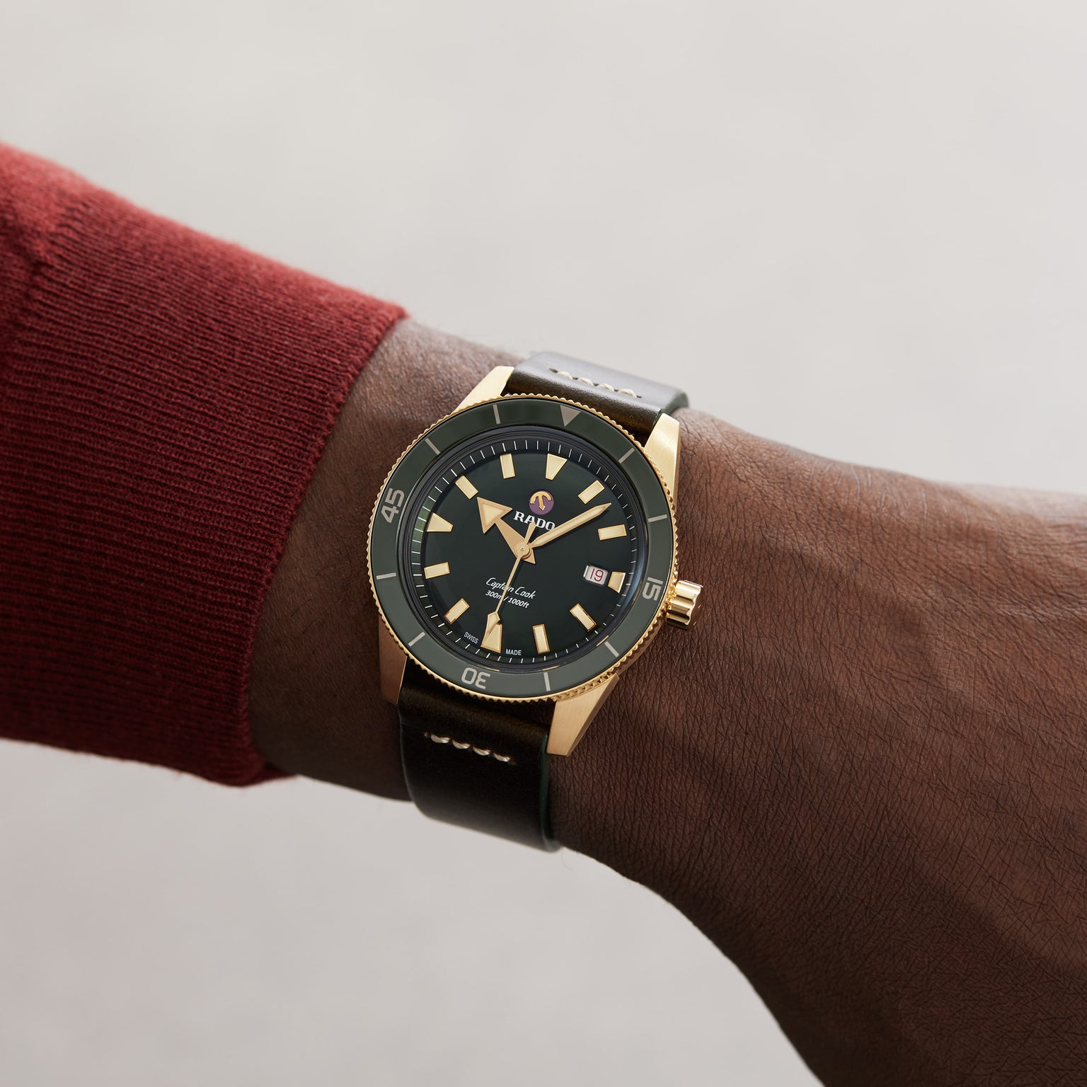 A person wearing a red sleeve sports the Rado Captain Cook Automatic Bronze, a gold and black wristwatch with a black leather strap.