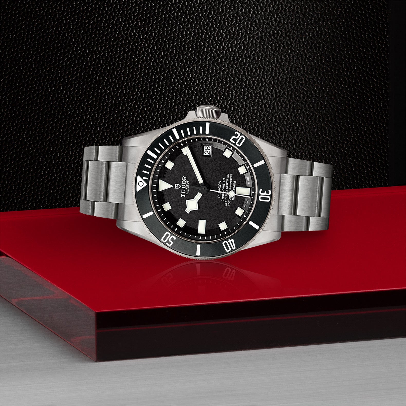 The TUDOR Pelagos, a stainless steel divers tool watch with a black dial and rotating bezel, is displayed on a red platform against a textured black background.