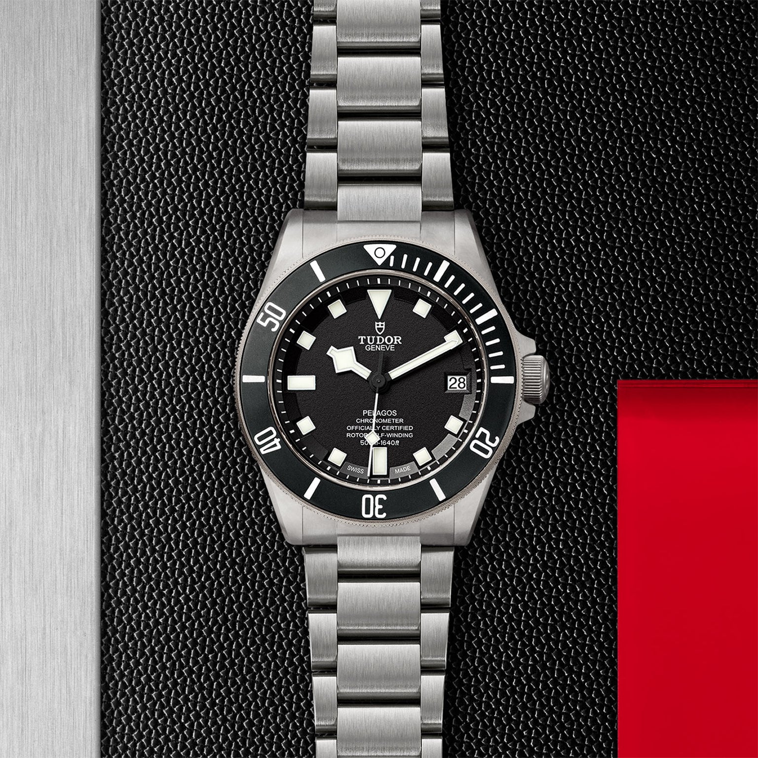 A TUDOR Pelagos stainless steel divers watch with a black bezel and dial is showcased on a textured black and metal background featuring a red accent.