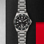 A TUDOR Pelagos stainless steel divers watch with a black bezel and dial is showcased on a textured black and metal background featuring a red accent.