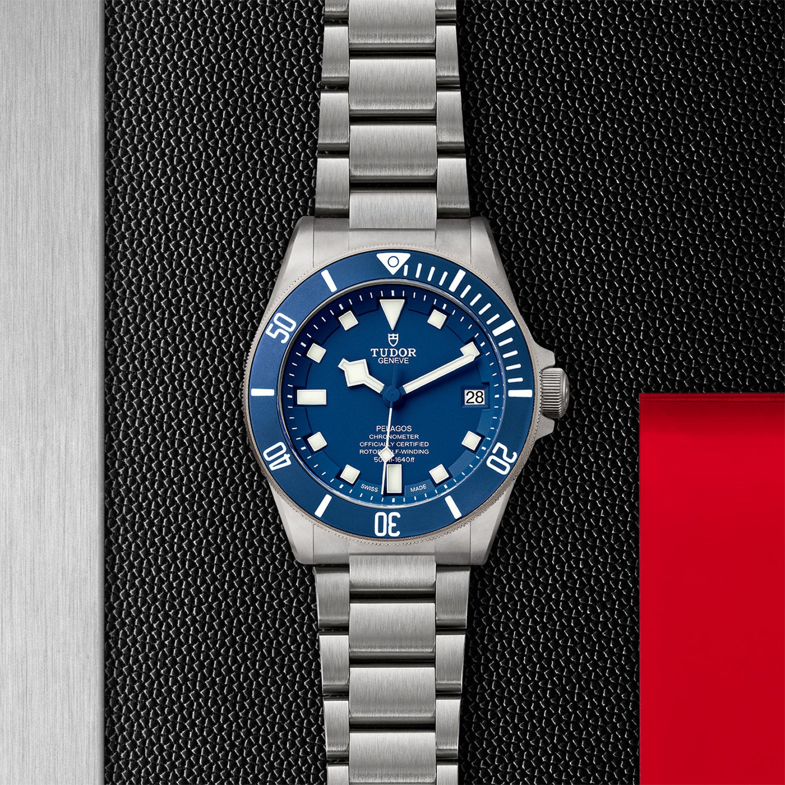 A TUDOR Pelagos wristwatch with a blue bezel and dial, silver hour markers, and date display rests on a textured black and metallic surface with a red accent.