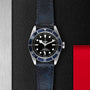 A TUDOR Black Bay watch with a black dial, blue bezel, and black leather strap is shown on a textured black surface accented with metal and red details.