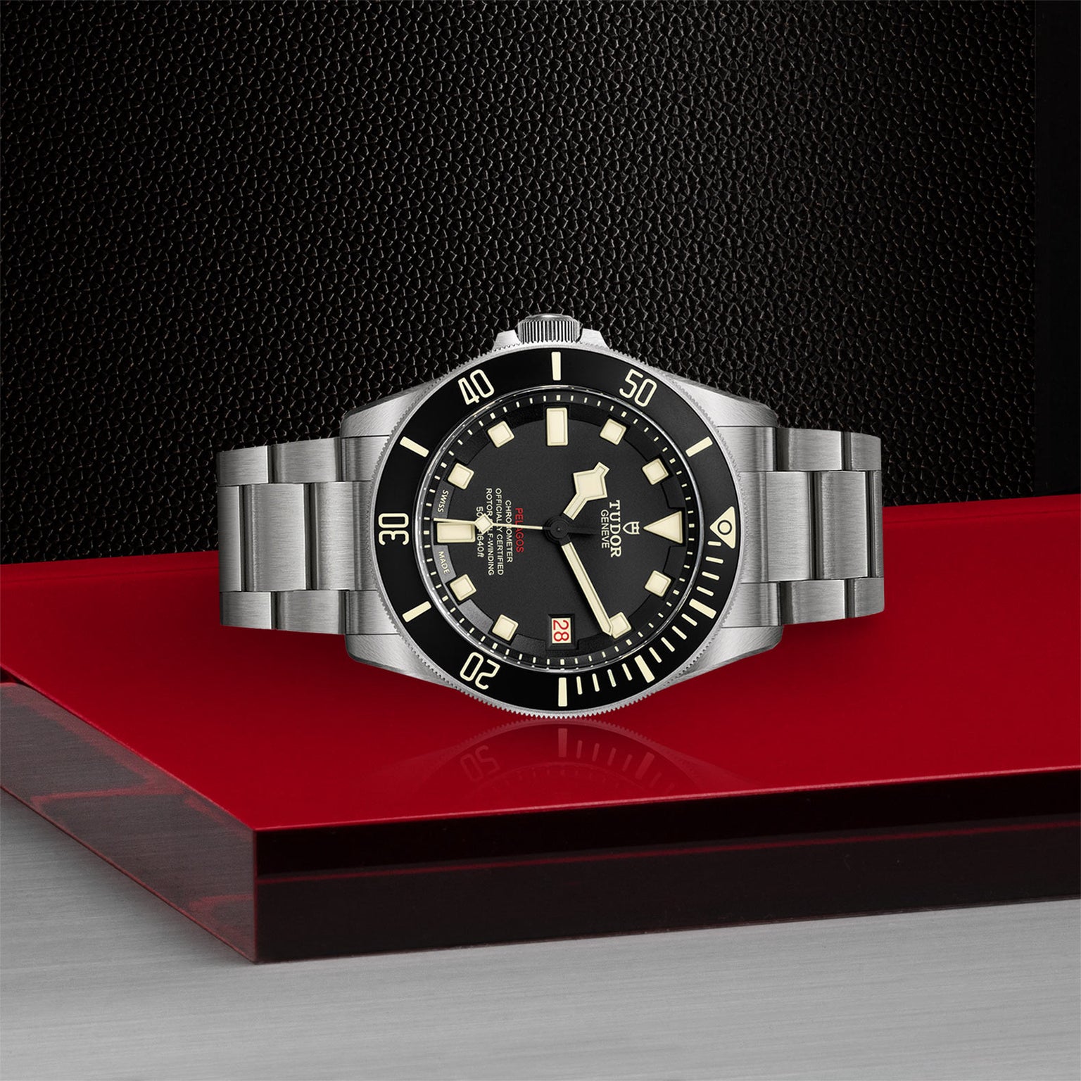 The TUDOR Pelagos LHD, a stainless steel wristwatch with a black dial and bezel, is displayed on a red and silver surface.