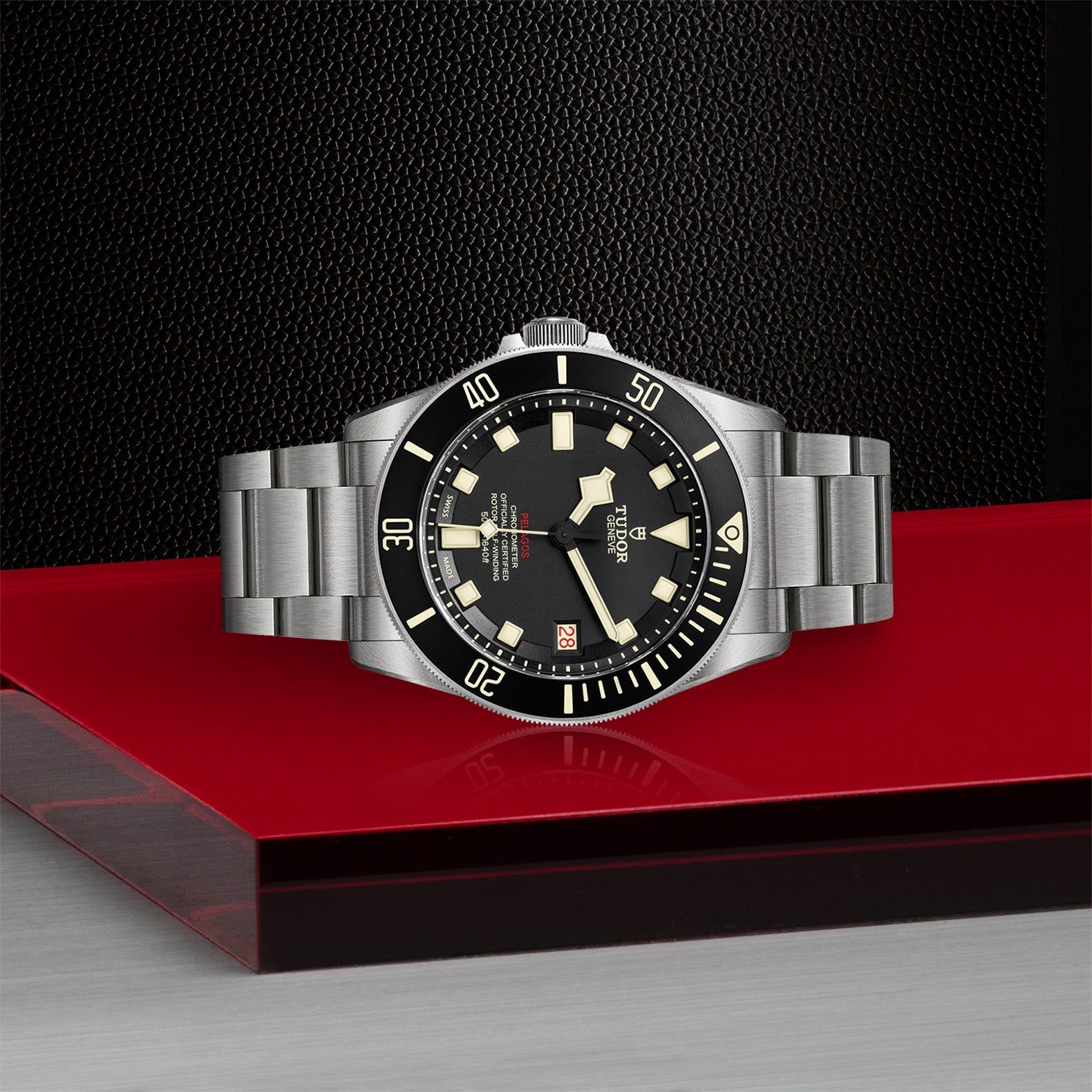 The TUDOR Pelagos LHD, a stainless steel wristwatch with a black dial and bezel, is displayed on a red and silver surface.