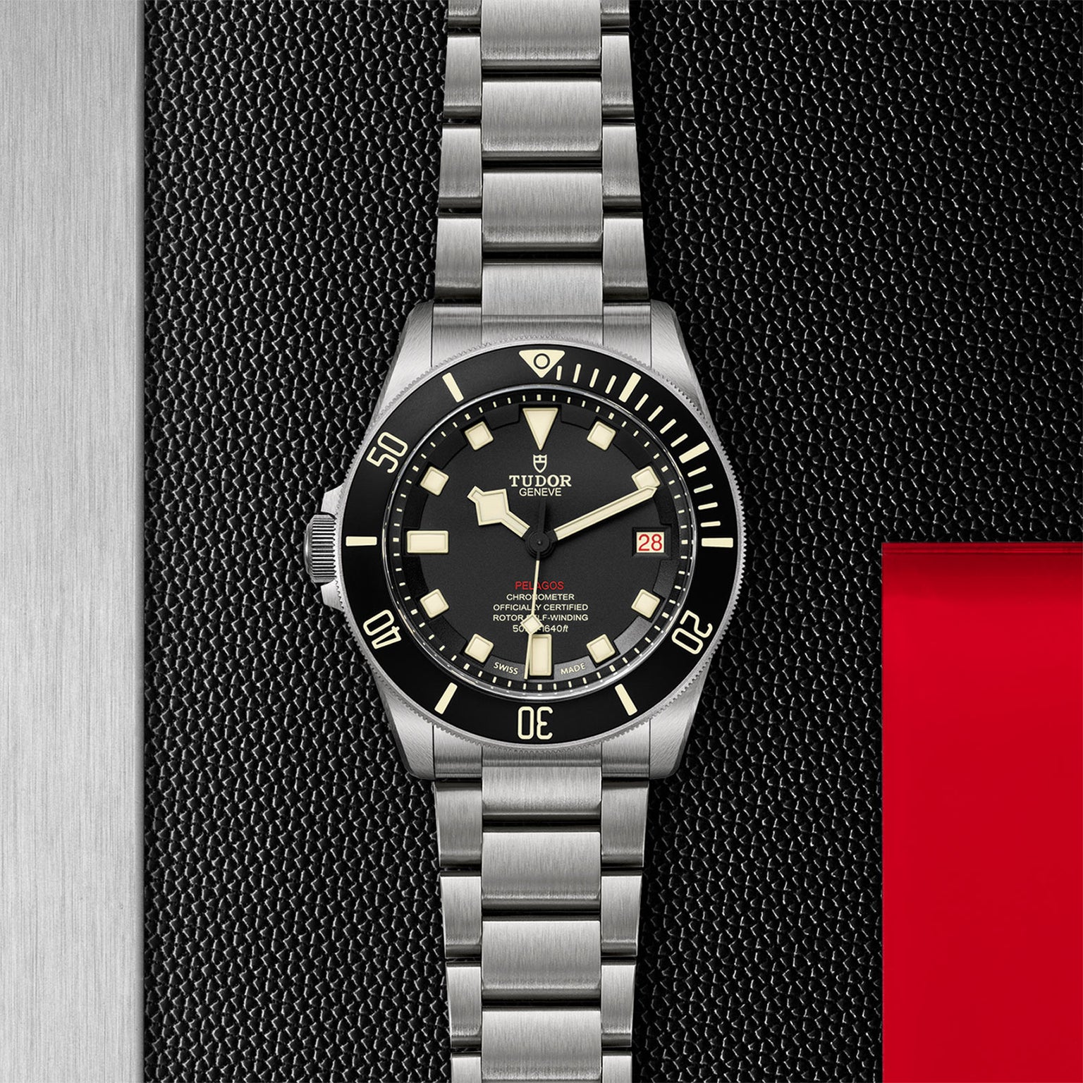 The TUDOR Pelagos LHD, a stainless steel diver’s watch with a black bezel, cream markers, and metal bracelet, is displayed against a textured black and red background.