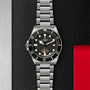 The TUDOR Pelagos LHD, a stainless steel diver’s watch with a black bezel, cream markers, and metal bracelet, is displayed against a textured black and red background.
