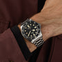 The TUDOR Pelagos LHD, a stainless steel diver’s watch with a black bezel and dial, is worn on the left wrist and partially concealed by a maroon coat sleeve.