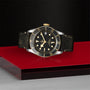 A TUDOR Black Bay S&G wristwatch with a black and gold finish and date display is showcased on a red platform against a textured black backdrop, highlighting its self-winding mechanical movement.