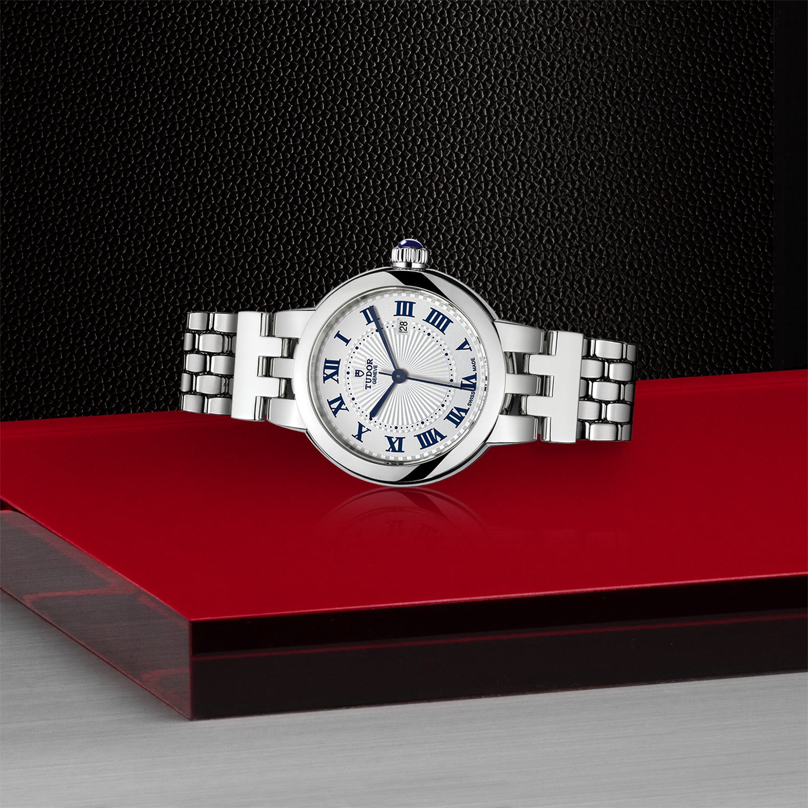 The TUDOR Clair de Rose, a silver luxury wristwatch with a white face and blue Roman numerals, is displayed on a red and black surface.