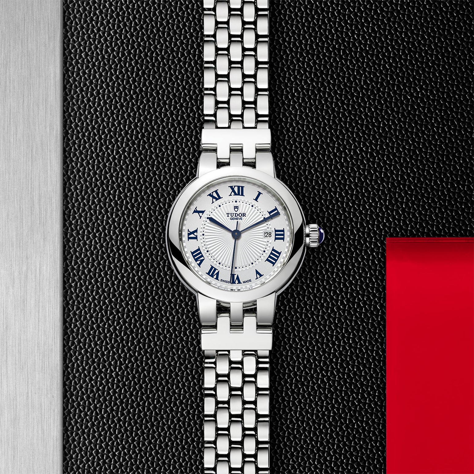 The TUDOR Clair de Rose features a silver linked bracelet, white dial, and blue Roman numerals set against a textured black background.