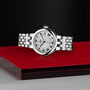 The TUDOR Clair de Rose silver wristwatch with Roman numerals rests on a red and gray surface, set against a black textured background.