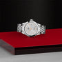 A TUDOR 1926 ladies wristwatch with a white textured dial and date display sits on a red platform against a dark, textured backdrop.
