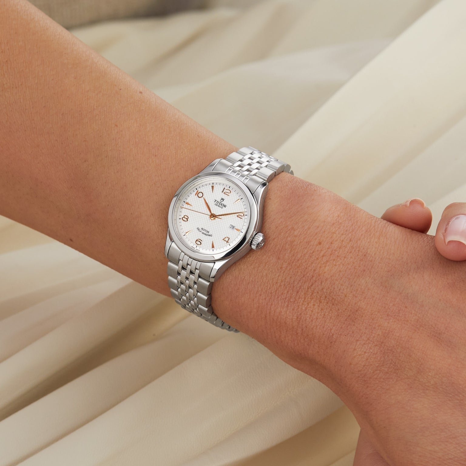 A TUDOR 1926 silver ladies’ wristwatch with a metal link band is shown on a person’s wrist against a beige fabric background.