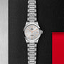 A silver TUDOR 1926 wristwatch with a metal link band and white textured dial is shown upright against a black and red textured background.