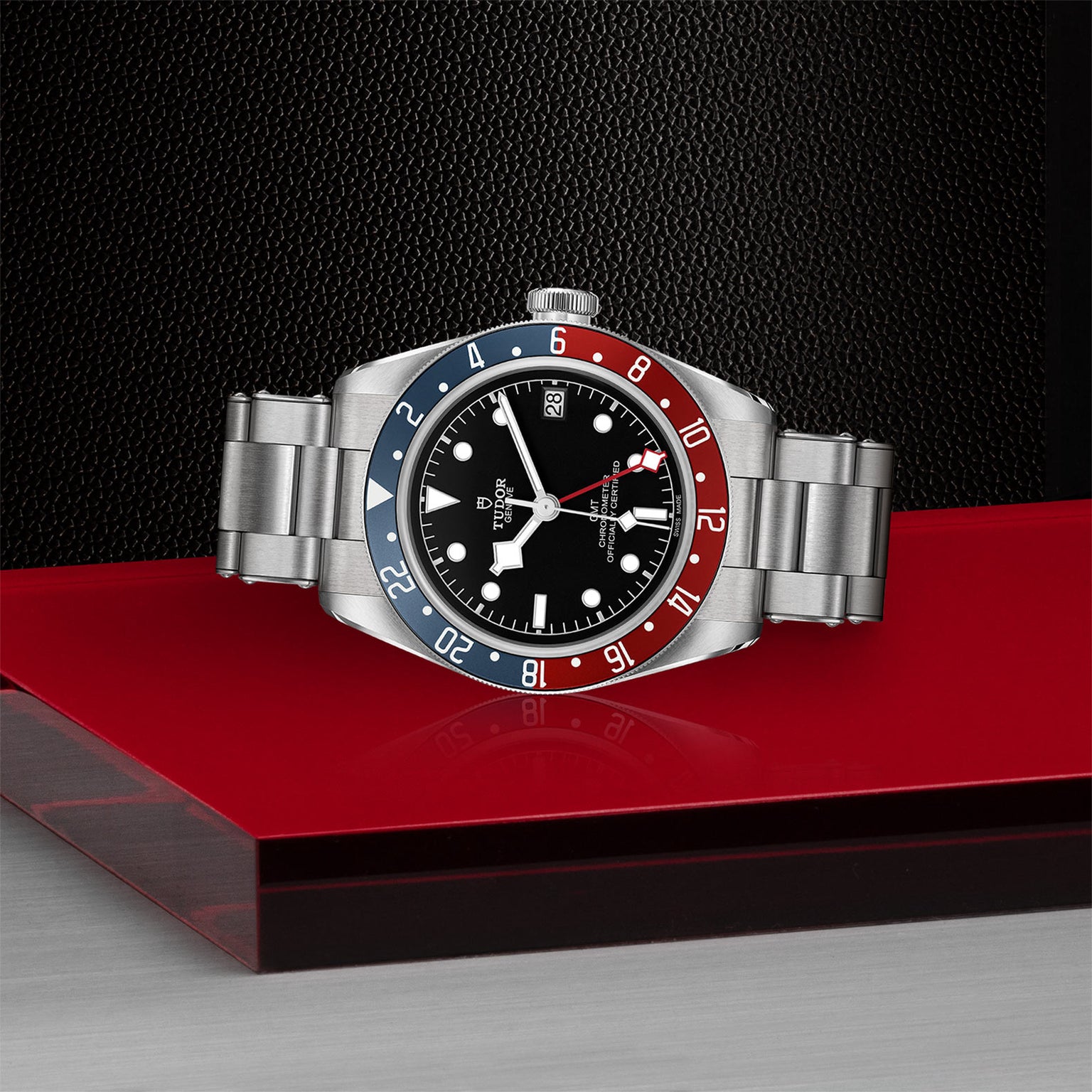 The TUDOR Black Bay GMT is a stainless steel wristwatch with a red and blue bezel, black dial, and metal bracelet displayed on a red surface.
