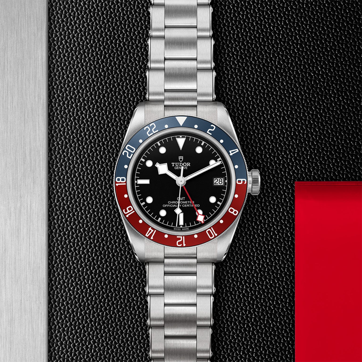 A TUDOR Black Bay GMT wristwatch with a stainless steel bracelet, black dial, and red-blue bezel is shown on a textured black and metallic surface.