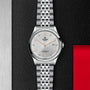 A TUDOR 1926 wristwatch with stainless steel bracelet, white dial, orange numerals, date display, and self-winding movement rests on a textured black background with metal and red accents.